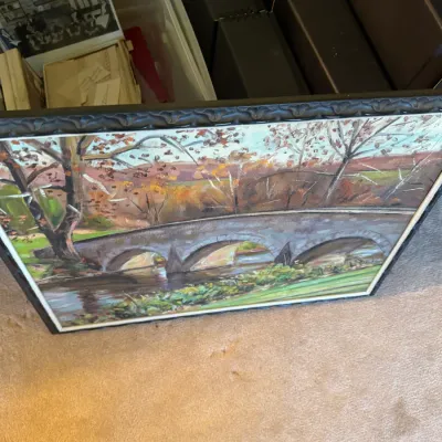 Sale Photo Thumbnail #339: Framed landscape oil painting depicting a stone arch bridge over a river in an autumn setting, stored alongside boxes of memorabilia.