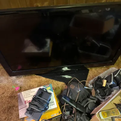 Sale Photo Thumbnail #99: Black flat-screen television sitting on a carpeted floor surrounded by various electronics, cables, power adapters, and camera bags.