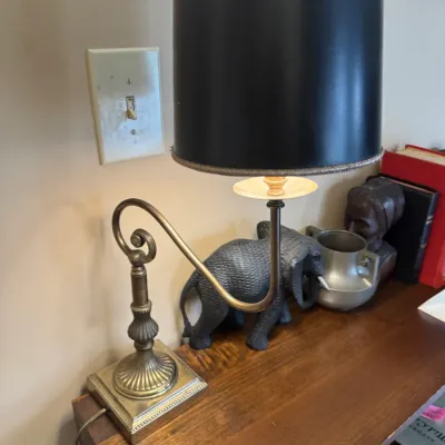 Sale Photo Thumbnail #317: Traditional brass gooseneck desk lamp with a tall black drum shade, set on a wooden tabletop near decorative figurines.