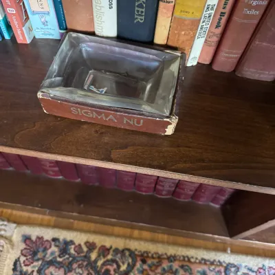 Sale Photo Thumbnail #309: Vintage Sigma Nu fraternity leather-wrapped glass ashtray sitting on a wooden bookshelf, showing some edge wear to the leather.