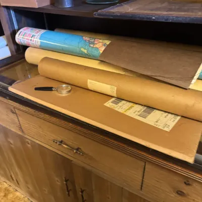 Sale Photo Thumbnail #94: Collection of rolled vintage wall maps and documents resting on a wooden cabinet alongside a small magnifying glass.