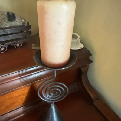 Sale Photo Thumbnail #323: Wrought iron spiral candlestick holder featuring a conical base and a large cream pillar candle, set on a wooden surface.