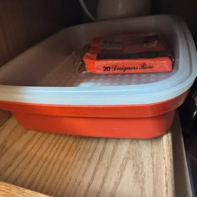 Sale Photo Thumbnail #228: Vintage orange Tupperware rectangular baking dish with a sheer lid, sitting inside a wooden cabinet.