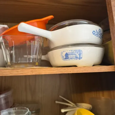 Sale Photo Thumbnail #258: Vintage Corning Ware skillet dishes featuring Cornflower and Homestead blue patterns, alongside clear glass bakeware and plastic kitchen utensils stored in a cabinet.