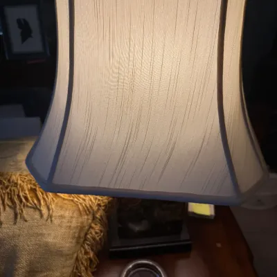 Sale Photo Thumbnail #350: Illuminated table lamp with a classic off-white pleated bell shade, shown alongside a silver-toned coaster on a wooden tabletop.