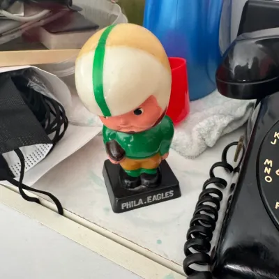 Sale Photo Thumbnail #26: Vintage Philadelphia Eagles bobblehead figure resting on a counter, featuring a green and gold uniform with a helmet, next to a black rotary phone.