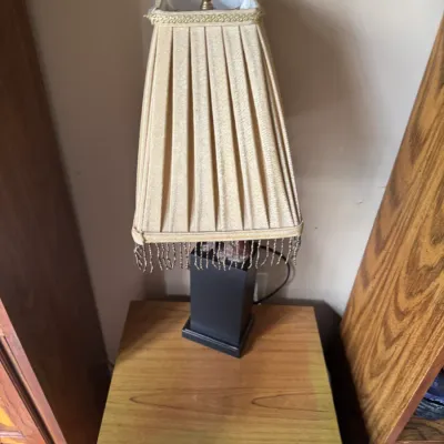 Sale Photo Thumbnail #301: Vintage table lamp featuring a dark rectangular pedestal base and a pleated gold fabric shade with beaded fringe trim.