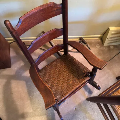 Sale Photo Thumbnail #345: Vintage child-sized wooden rocking chair featuring a ladder-back frame and a woven reed seat, showing visible wear and minor seat damage.