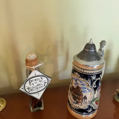 Sale Photo Thumbnail #326: German-style ceramic beer stein with a pewter lid alongside a small decorative glass bottle on a wooden surface.