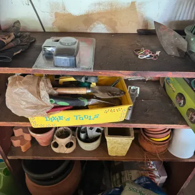 Sale Photo Thumbnail #193: Wooden workshop shelves filled with gardening supplies, hand tools in a yellow bin, terra cotta pots, and bags of peat moss.