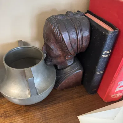 Sale Photo Thumbnail #315: Hand-carved wooden bust bookend alongside a vintage metal pot vessel and hardcover books.