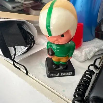 Sale Photo Thumbnail #208: Vintage Philadelphia Eagles papier-mâché or composition bobblehead figure from the 1960s, featuring a square black base and retro helmet design.