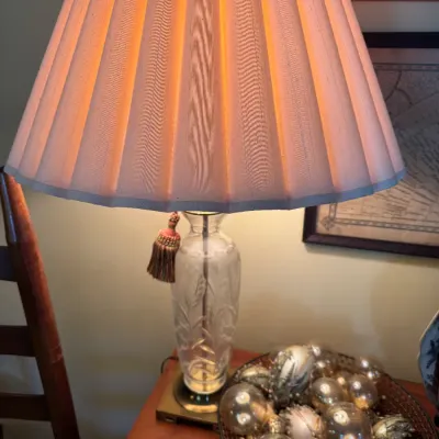 Sale Photo Thumbnail #343: Cut crystal table lamp with a pleated fabric shade and tassel detail, resting on a wooden side table alongside a decorative tray of glass ornaments.