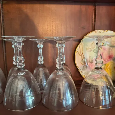 Sale Photo Thumbnail #396: Set of clear crystal stemmed wine glasses with subtle etched floral details, stored upside down on a wooden cabinet shelf.