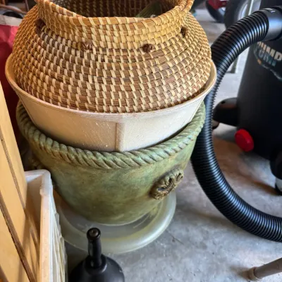 Sale Photo Thumbnail #207: A stack of decorative home goods including a hand-woven basket, a fabric lampshade, and a green rope-rimmed planter pot, shown with a hand bell in the foreground.