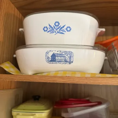 Sale Photo Thumbnail #259: Vintage CorningWare casserole dishes featuring Blue Cornflower and Homestead patterns with glass lids inside a wooden cabinet.