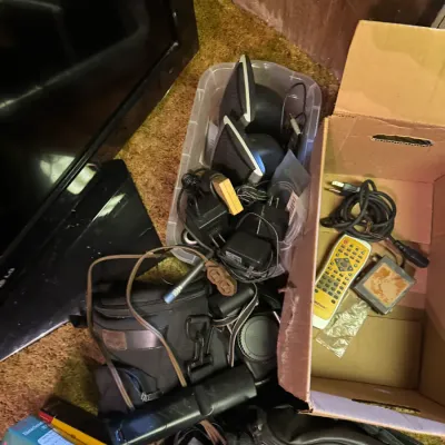 Sale Photo Thumbnail #98: Collection of assorted electronics and accessories, including desktop computer speakers, power adapters, camera bags, and a television remote stored in plastic and cardboard containers.