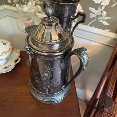 Sale Photo Thumbnail #389: Antique Victorian silver-plated insulated water pitcher with engraved floral designs, displayed on a wooden surface alongside a matching trophy cup.