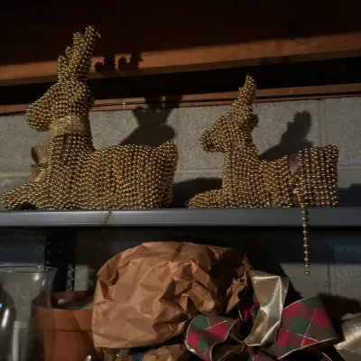 Sale Photo Thumbnail #70: Pair of gold bead-covered reclining reindeer Christmas tabletop decorations resting on a shelf above plaid holiday bows.
