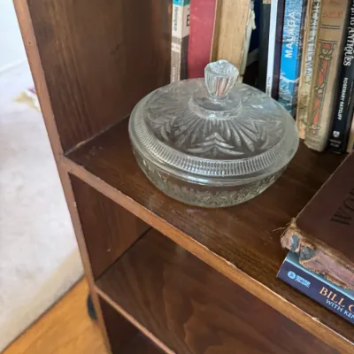 Sale Photo Thumbnail #310: Vintage pressed clear glass covered candy dish or powder jar with a matching lid, displayed on a wooden shelf beside books.