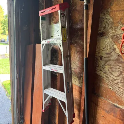 Sale Photo Thumbnail #191: Werner aluminum stepladder stored in a garage alongside a long-handled lawn rake and wood planks.