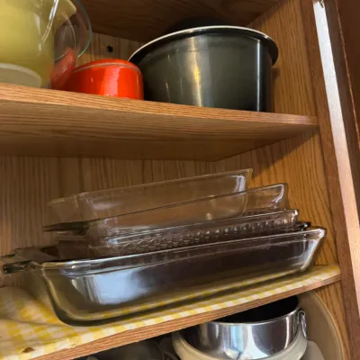 Sale Photo Thumbnail #255: A wooden kitchen cabinet filled with assorted glassware and cookware, including stacked clear Pyrex baking dishes, vintage nesting mixing bowls, and a crock pot liner.