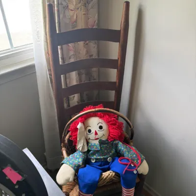 Sale Photo Thumbnail #105: Vintage Raggedy Andy rag doll seated in a woven basket chair, set against a tall dark wood ladder-back dining chair.