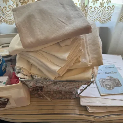 Sale Photo Thumbnail #167: Stack of folded beige and cream fabrics resting in a wire mesh basket on an ironing board.