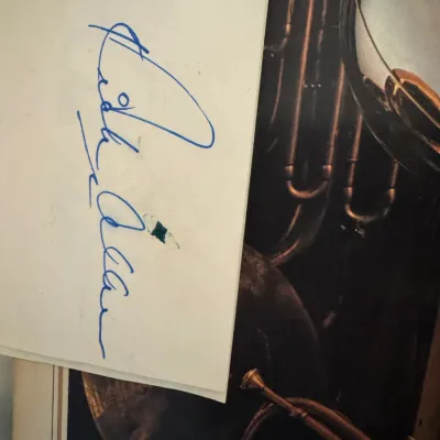 Sale Photo Thumbnail #159: Hand-signed signature on paper in blue ink, laid over a framed brass musical instrument artwork.