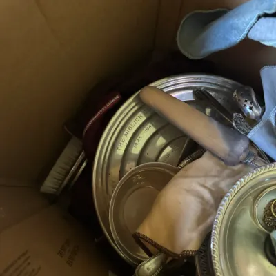 Sale Photo Thumbnail #358: Cardboard box containing a lot of vintage silverplate and metalware, including trophy platters, serving pieces, and brushes.
