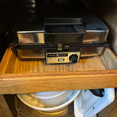 Sale Photo Thumbnail #251: Vintage General Electric chrome grill and waffle baker stored on a wooden cabinet shelf.