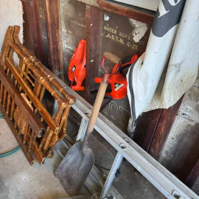 Sale Photo Thumbnail #204: A metal shovel with a wooden handle resting against an aluminum ladder, surrounded by vintage wooden chair frames and Black & Decker power tools in a garage.