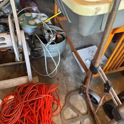 Sale Photo Thumbnail #195: Assorted workshop and utility items, including a long coiled orange extension cord, a metal hand truck, and various small appliances stored on a concrete floor.