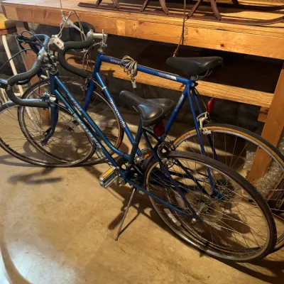 Sale Photo Thumbnail #78: Two vintage blue 10-speed road bikes, including a Panasonic Sport 500, parked in a basement workshop near a wooden workbench.
