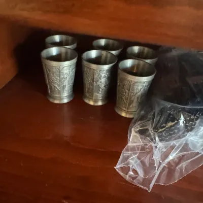 Sale Photo Thumbnail #404: Set of six vintage pewter shot glasses featuring embossed relief scenes, displayed on a wooden shelf.