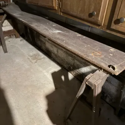 Sale Photo Thumbnail #71: Rustic long wooden workbench supported by saw horses, showing age, dust, and heavy surface wear in a workshop environment.
