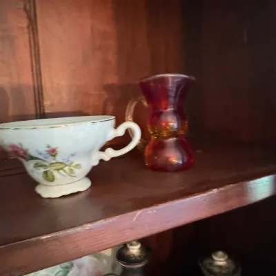 Sale Photo Thumbnail #394: Vintage porcelain teacup with floral motifs alongside a small ruby and amber glass pitcher, displayed on a wooden shelf.