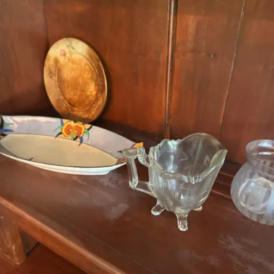 Sale Photo Thumbnail #400: Collection of vintage glassware and ceramics displayed on a wooden hutch shelf, including a clear pressed glass creamer with footed base and an Art Deco hand-painted dish.