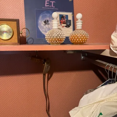 Sale Photo Thumbnail #149: Display shelf featuring a Michael Jackson E.T. soundtrack record, a pair of vintage Fenton hobnail glass perfume bottles, and a small framed weather instrument.