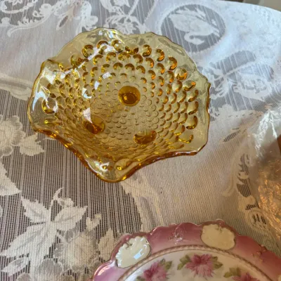 Sale Photo Thumbnail #414: Vintage amber hobnail glass three-footed candy dish with a ruffled edge, displayed alongside a floral porcelain plate on a lace tablecloth.