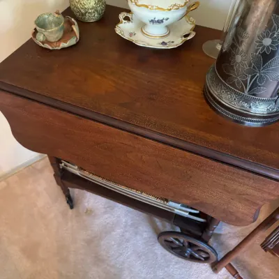 Sale Photo Thumbnail #386: Vintage mahogany drop-leaf tea cart on spoked wheels, displayed with decorative metal pitchers and floral porcelain tableware.