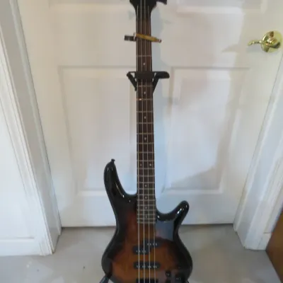 Sale Photo Thumbnail #98: Ibanez 4-string electric bass guitar with a sunburst finish, displayed on a black folding stand.
