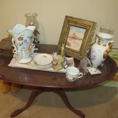Sale Photo Thumbnail #72: Collection of decorative vintage items including floral porcelain vases, teacups, glass hurricane lamps, and framed art atop an oval mahogany accent table with carved legs.