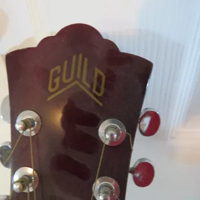 Sale Photo Thumbnail #108: Headstock of a Guild acoustic guitar featuring gold inlay logo and chrome tuning pegs.