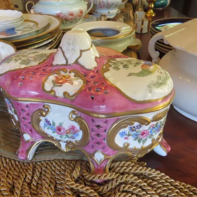 Sale Photo Thumbnail #23: Hand-painted porcelain footed trinket box with pink glaze, reticulated lattice work, floral panels, and gilt trim, shown alongside other vintage tableware.