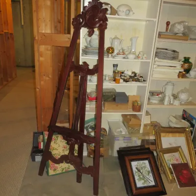 Sale Photo Thumbnail #62: Ornate carved dark wood floor easel standing in front of shelving units filled with ceramics, dishware, and framed artwork.