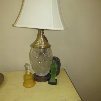 Sale Photo Thumbnail #89: Vintage cut glass table lamp with brass accents and a white fabric shade, displayed alongside an amber glass bell and a small green figurine.