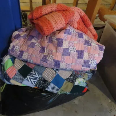 Sale Photo Thumbnail #50: Stack of handmade quilts and a pink and coral crocheted blanket, resting atop a black plastic trash bag.