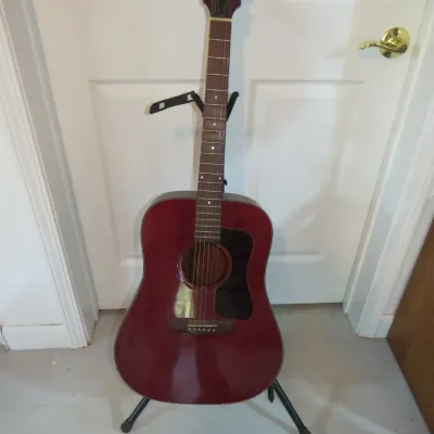 Sale Photo Thumbnail #107: Burgundy acoustic dreadnought guitar resting on a black folding tripod stand.