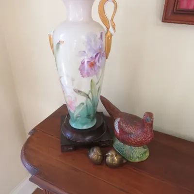 Sale Photo Thumbnail #16: Hand-painted porcelain urn with floral motif and gilded handles mounted on a wooden base, displayed alongside a ceramic pheasant figurine and two small brass bird statues.